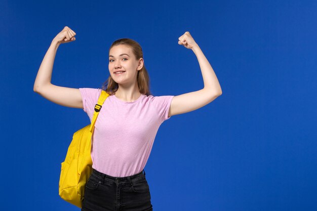 Vista frontal de la estudiante en camiseta rosa con mochila amarilla sonriendo y flexionando en la pared azul claro