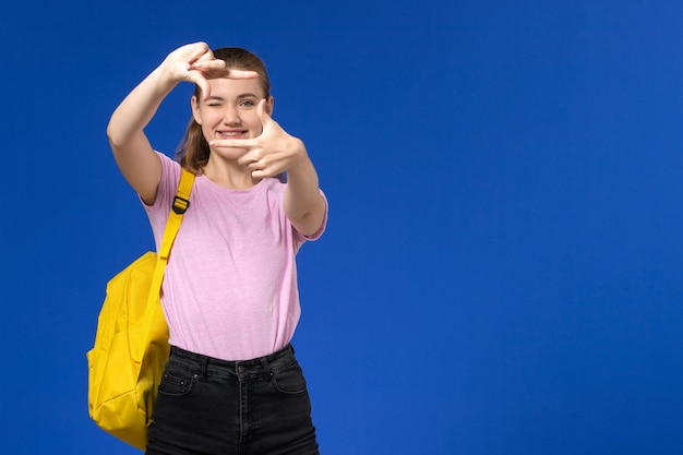 Vista frontal de la estudiante en camiseta rosa con mochila amarilla sonriendo en la pared azul claro