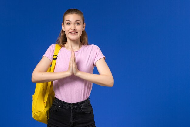 Vista frontal de la estudiante en camiseta rosa con mochila amarilla sonriendo en la pared azul claro