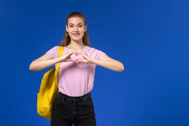 Vista frontal de la estudiante en camiseta rosa con mochila amarilla sonriendo en la pared azul