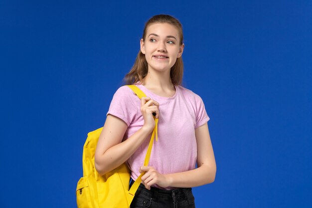 Vista frontal de la estudiante en camiseta rosa con mochila amarilla sonriendo en la pared azul