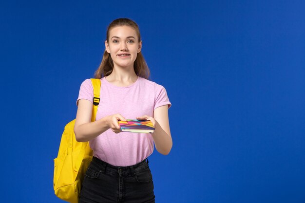 Vista frontal de la estudiante en camiseta rosa con mochila amarilla sosteniendo un cuaderno en la pared azul