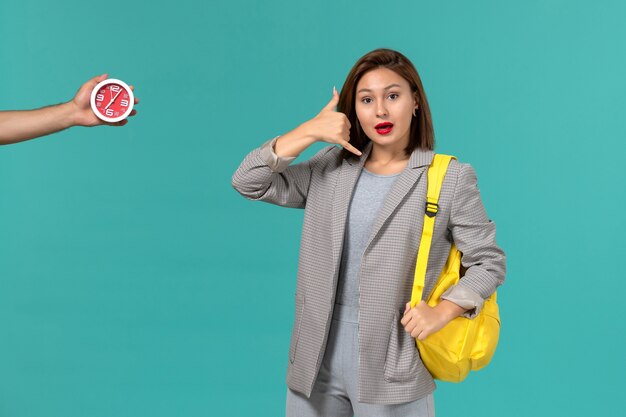 Vista frontal de la estudiante en chaqueta gris con mochila amarilla en la pared azul claro