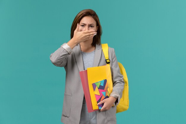 Vista frontal de la estudiante en chaqueta gris con mochila amarilla sosteniendo archivos y cuaderno cubriendo su boca en la pared azul