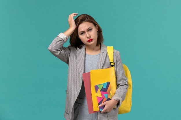 Vista frontal de la estudiante en chaqueta gris con mochila amarilla sosteniendo archivos y cuaderno con dolor de cabeza en la pared azul