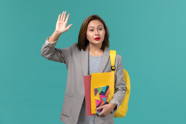 Vista frontal de la estudiante en chaqueta gris con mochila amarilla sosteniendo archivos y cuaderno ondeando en la pared azul