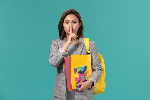 Vista frontal de la estudiante en chaqueta gris con mochila amarilla sosteniendo archivos y cuaderno en la pared azul