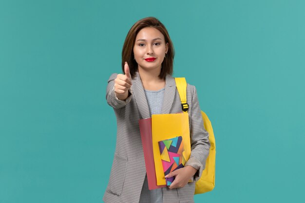 Vista frontal de la estudiante en chaqueta gris con mochila amarilla sosteniendo archivos y cuaderno sonriendo en la pared azul