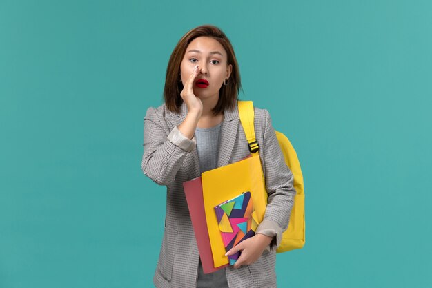 Vista frontal de la estudiante en chaqueta gris con mochila amarilla sosteniendo archivos y cuaderno susurrando en la pared azul