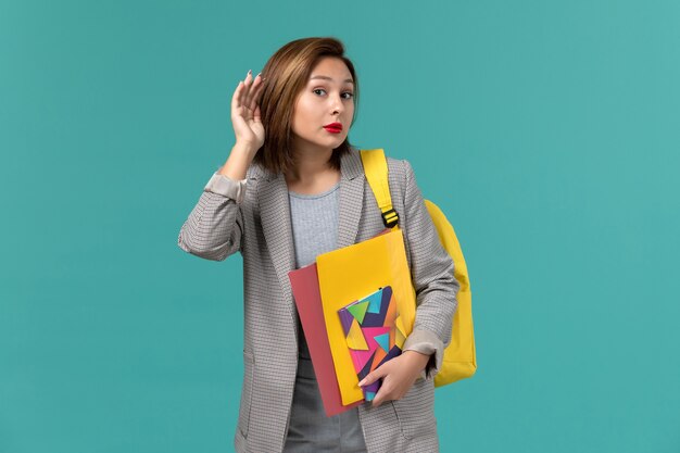 Vista frontal de la estudiante en chaqueta gris con mochila amarilla sosteniendo archivos y cuaderno tratando de escuchar en la pared azul