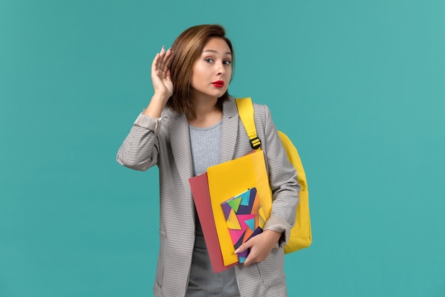 Vista frontal de la estudiante en chaqueta gris con mochila amarilla sosteniendo archivos y cuaderno tratando de escuchar en la pared azul
