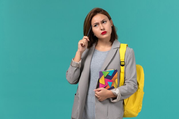 Vista frontal de la estudiante en chaqueta gris con mochila amarilla sosteniendo el cuaderno en la pared azul