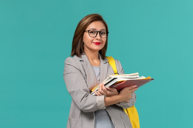 Vista frontal de la estudiante en chaqueta gris mochila amarilla sosteniendo libros en la pared azul claro