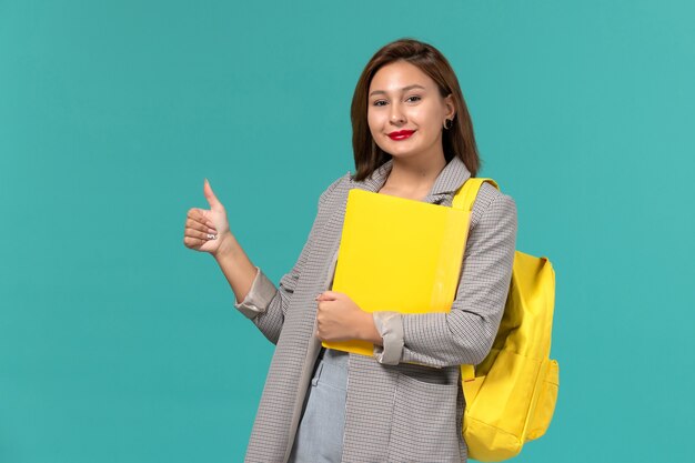 Vista frontal de la estudiante en chaqueta gris con su mochila amarilla y sosteniendo archivos en la pared azul claro