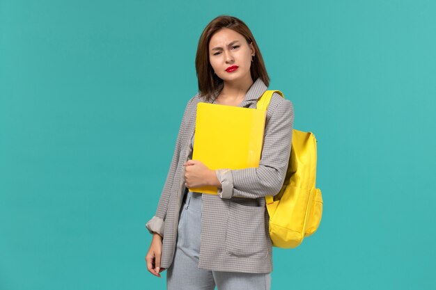 Vista frontal de la estudiante en chaqueta gris con su mochila amarilla y sosteniendo archivos en la pared azul claro