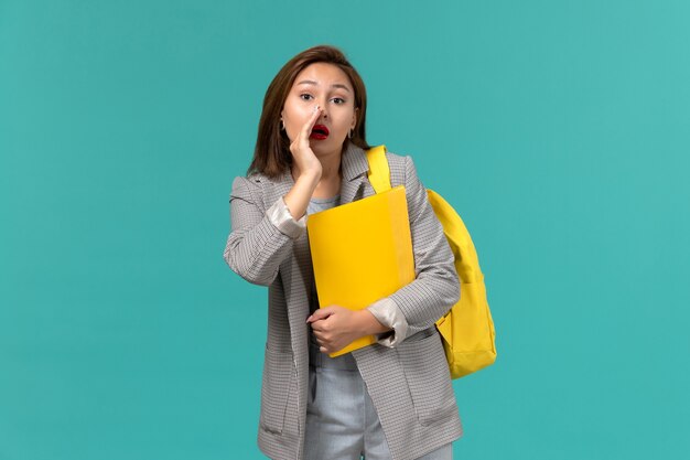 Vista frontal de la estudiante en chaqueta gris con su mochila amarilla y sosteniendo archivos susurrando en la pared azul claro