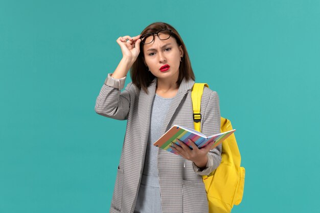 Vista frontal de la estudiante en chaqueta gris con su mochila amarilla sosteniendo el cuaderno en la pared azul claro
