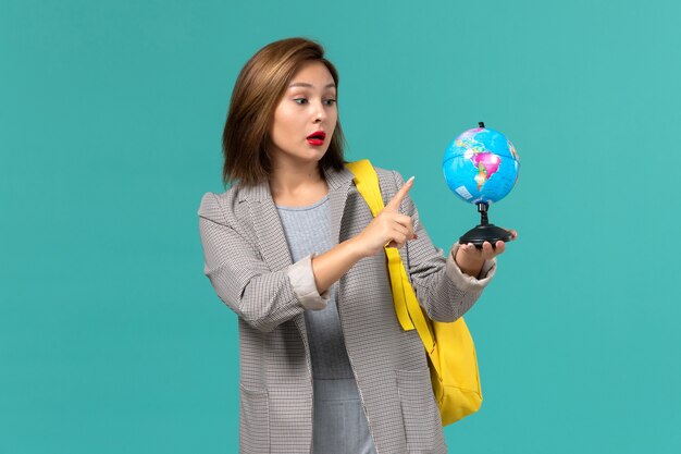 Vista frontal de la estudiante en chaqueta gris con su mochila amarilla sosteniendo un pequeño globo en la pared azul claro