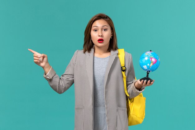 Vista frontal de la estudiante en chaqueta gris con su mochila amarilla sosteniendo un pequeño globo en la pared azul claro