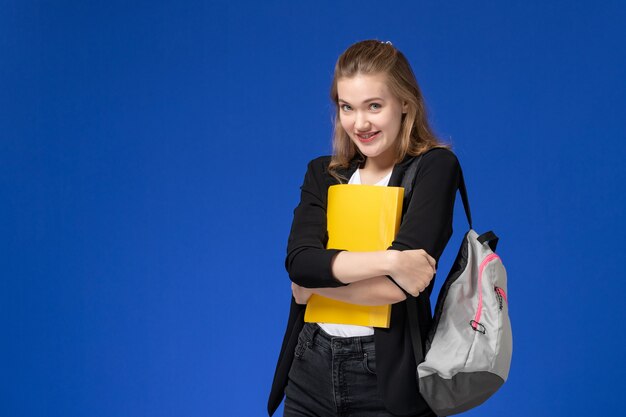 Vista frontal estudiante en chaqueta negra con mochila y sosteniendo un archivo amarillo en la lección de la universidad de la escuela de la pared azul