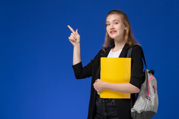 Vista frontal estudiante de chaqueta negra con mochila sosteniendo archivos amarillos en el escritorio azul lección de la universidad de la escuela de la escuela