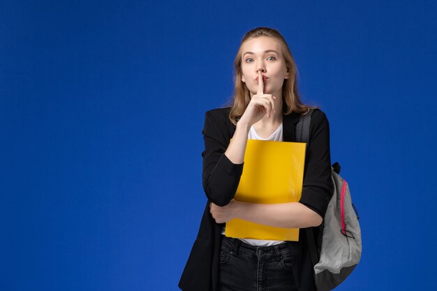 Vista frontal estudiante en chaqueta negra con mochila sosteniendo archivos amarillos en la lección de la universidad de la escuela de la pared azul