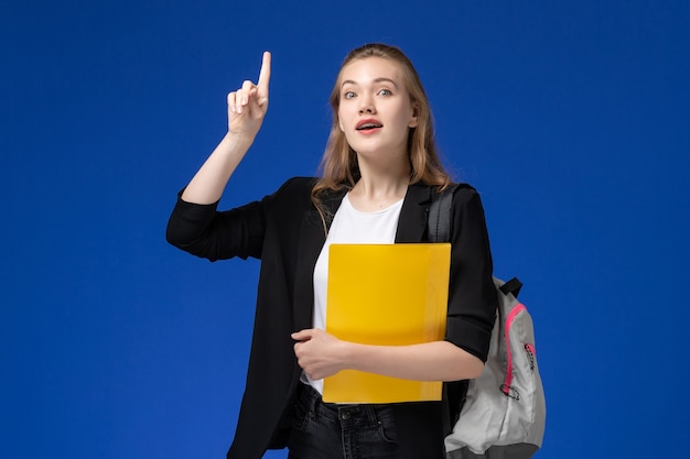 Vista frontal estudiante en chaqueta negra con mochila sosteniendo archivos amarillos en lecciones de la universidad de la escuela de la pared azul