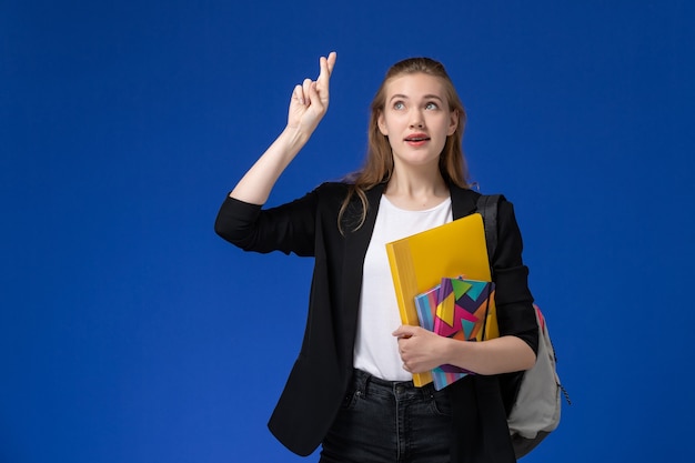 Vista frontal estudiante en chaqueta negra con mochila sosteniendo archivos con cuadernos cruzando los dedos sobre la pared azul lección universitaria de la universidad