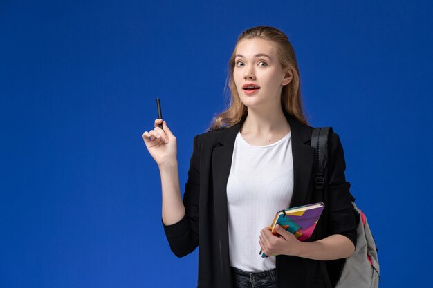 Vista frontal estudiante en chaqueta negra con mochila sosteniendo bolígrafo y cuaderno sobre pared azul lección universitaria