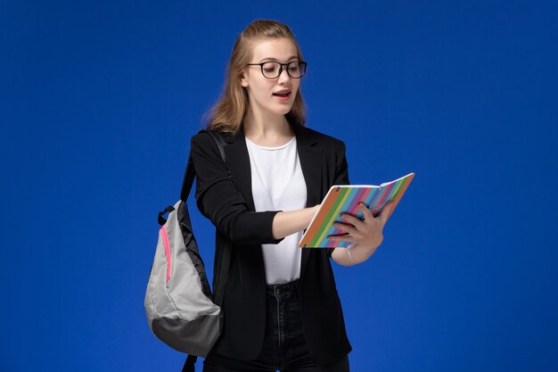 Vista frontal estudiante en chaqueta negra con mochila sosteniendo cuaderno y leyendo en la pared azul lecciones escuela colegio universidad