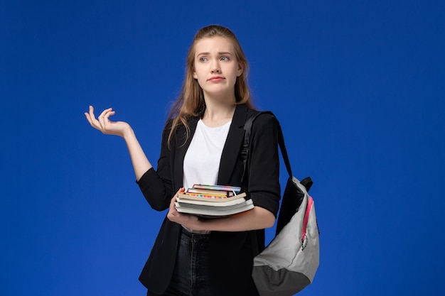 Vista frontal estudiante en chaqueta negra con mochila sosteniendo libros sobre la pared azul lección universitaria de la universidad