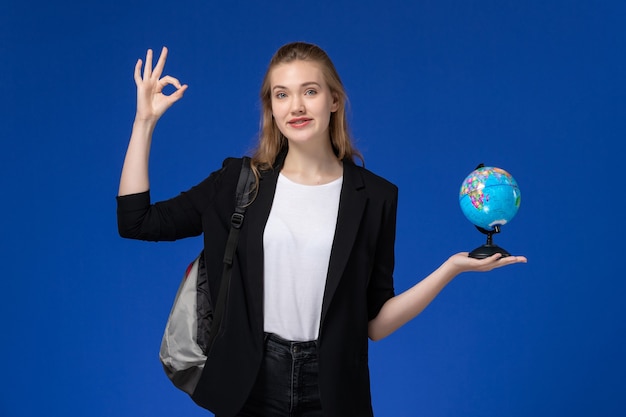 Vista frontal estudiante de chaqueta negra con mochila sosteniendo un pequeño globo en la pared azul claro escuela colegio universidad tierra