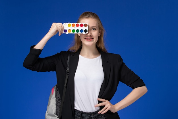 Foto gratuita vista frontal estudiante en chaqueta negra con mochila sosteniendo pinturas para dibujar en la pared azul dibujo art college university