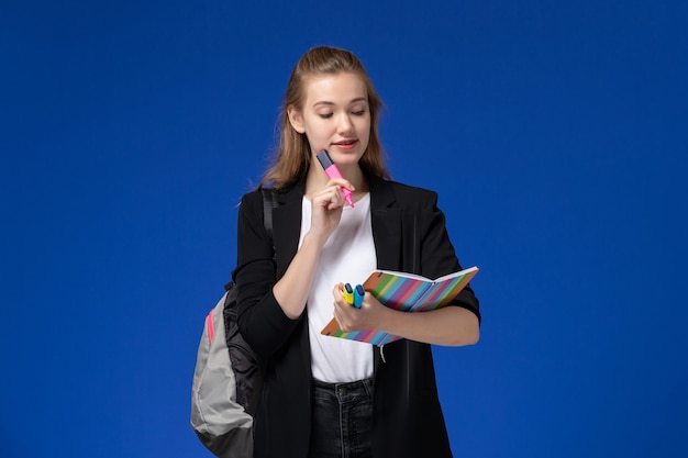 Vista frontal estudiante en chaqueta negra con mochila sosteniendo rotuladores y cuaderno en la pared azul lecciones escuela colegio universidad