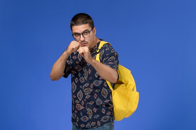 Vista frontal del estudiante masculino en camisa de algodón oscuro con mochila amarilla pose de boxeador en la pared azul