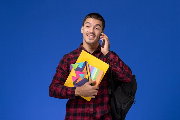 Vista frontal del estudiante masculino en camisa a cuadros roja con mochila con cuaderno y archivos hablando por teléfono en la pared azul