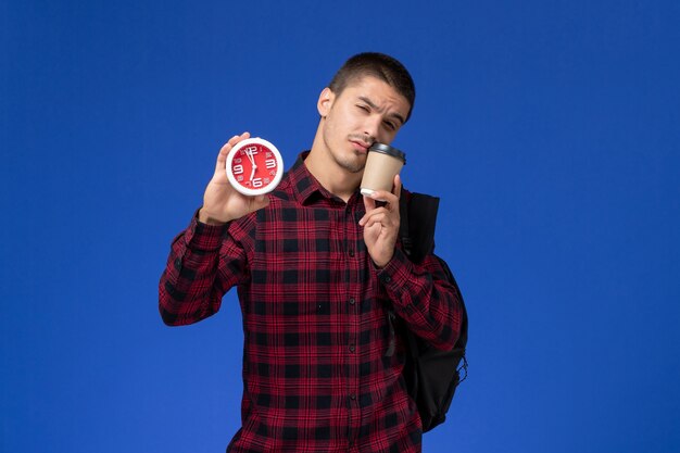 Vista frontal del estudiante masculino en camisa a cuadros roja con mochila sosteniendo relojes y café en la pared azul