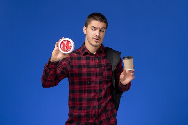 Vista frontal del estudiante masculino en camisa a cuadros roja con mochila sosteniendo relojes y café en la pared azul