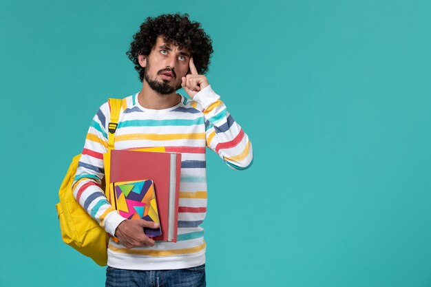 Vista frontal del estudiante masculino en camisa a rayas de colores con mochila amarilla sosteniendo archivos y cuadernos pensando en la pared azul