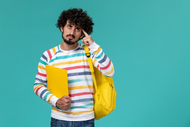 Vista frontal del estudiante masculino en camisa a rayas con mochila amarilla sosteniendo archivos pensando en la pared azul