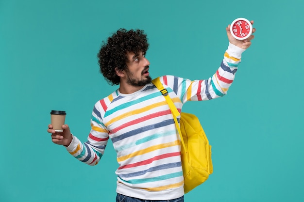 Vista frontal del estudiante masculino en camisa a rayas con mochila amarilla sosteniendo café y relojes en la pared azul