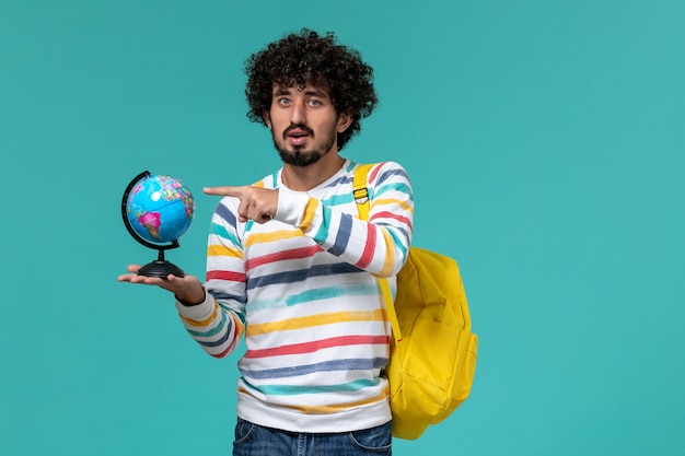 Vista frontal del estudiante masculino en camisa a rayas con mochila amarilla sosteniendo un pequeño globo redondo en la pared azul