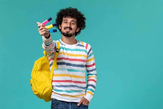 Vista frontal del estudiante masculino en camisa a rayas con mochila amarilla sosteniendo rotuladores de colores en la pared azul