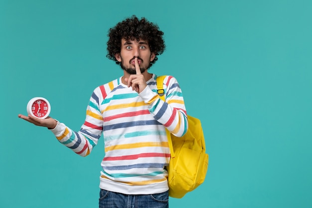 Vista frontal del estudiante masculino en camisa a rayas vistiendo una mochila amarilla sosteniendo relojes mostrando el signo de silencio en la pared azul