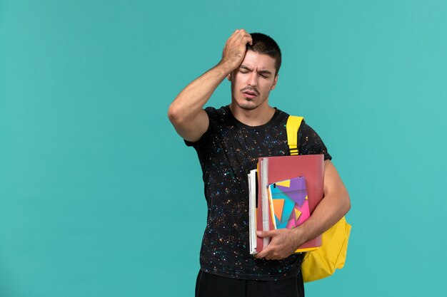 Vista frontal del estudiante masculino en camiseta oscura con mochila amarilla con cuaderno y archivos con dolor de cabeza en la pared azul