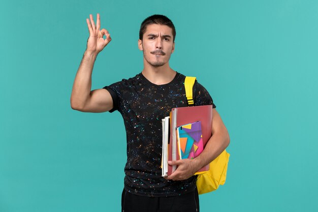 Vista frontal del estudiante masculino en camiseta oscura con mochila amarilla con cuaderno y archivos en la pared azul
