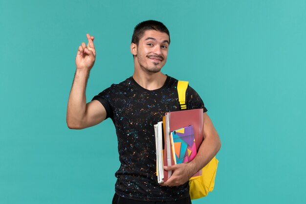 Vista frontal del estudiante masculino en camiseta oscura con mochila amarilla sosteniendo cuaderno y archivos cruzando los dedos sobre la pared azul