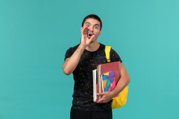 Vista frontal del estudiante masculino en camiseta oscura con mochila amarilla sosteniendo un cuaderno y archivos llamando a la pared azul