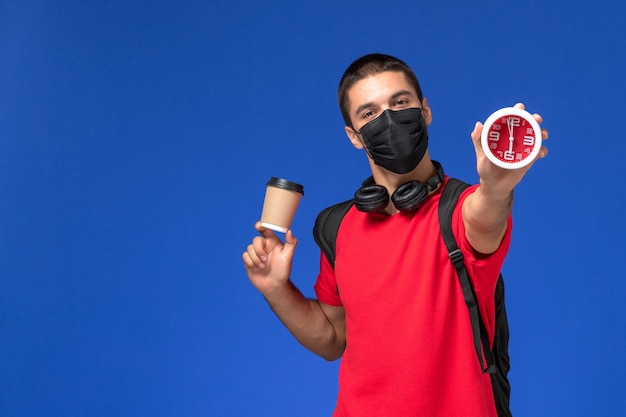 Vista frontal estudiante masculino en camiseta roja con máscara con mochila sosteniendo relojes café en el escritorio azul.