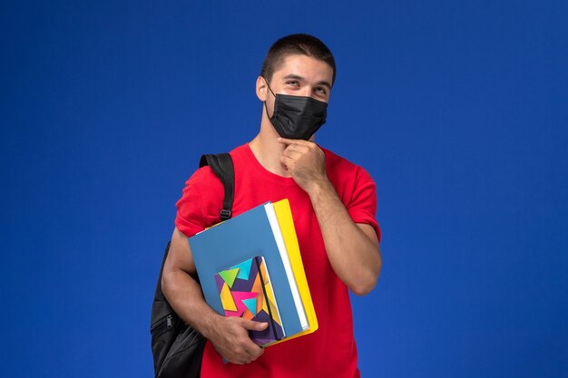 Vista frontal estudiante masculino en camiseta roja con mochila en máscara estéril negra sosteniendo cuadernos pensando en el fondo azul.